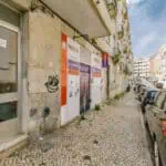 Stokarea - Mini Armazens Self storage Rua estreita de paralelepípedos ladeada por carros estacionados e prédios de apartamentos. Uma fachada de loja com sinalização e grafite é visível à esquerda. O céu está limpo e azul. Lidar em Portugal