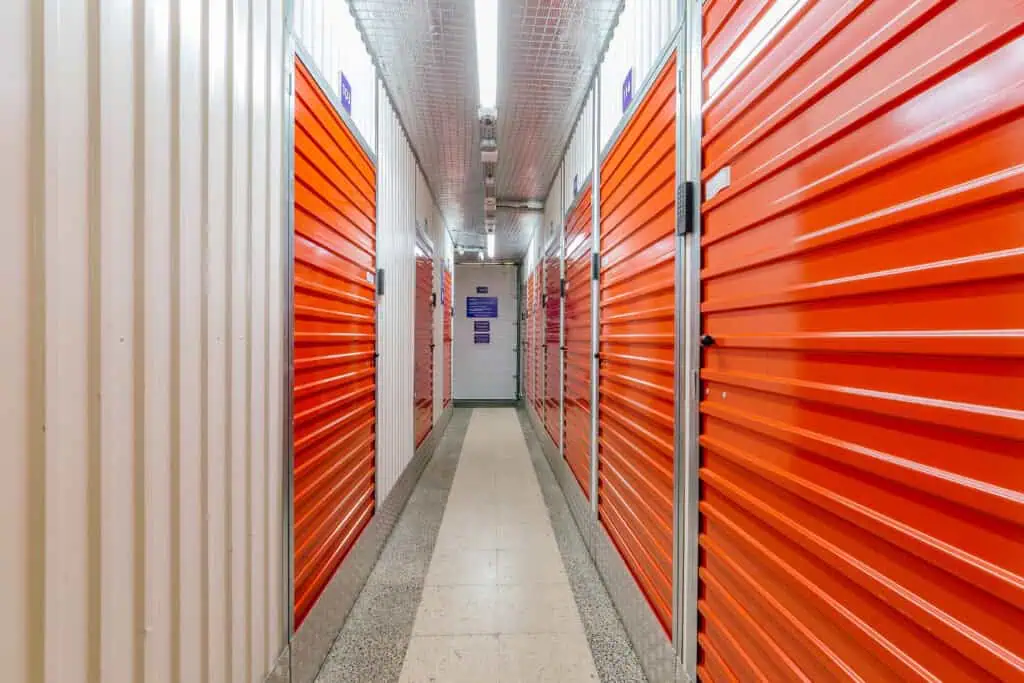 Stokarea - Mini Armazens Self storage Um corredor interno bem iluminado com portas de unidade de armazenamento laranja em ambos os lados. As paredes são de cor clara, e o piso do corredor é de ladrilho. Luzes suspensas correm ao longo do centro do teto, criando uma aparência limpa e organizada. Lidar em Portugal
