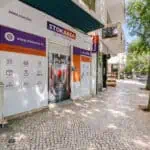 Stokarea - Mini Armazens Self storage Vista da rua de uma instalação de autoarmazenamento da Stokarea com uma placa branca e roxa. A entrada fica em uma calçada de ladrilhos, cercada por árvores e carros estacionados. A fachada da loja apresenta ícones e texto promovendo soluções de armazenamento. Lidar em Portugal