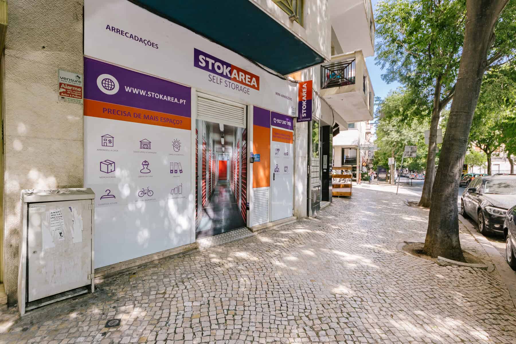 Stokarea - Mini Armazens Self storage Vista da rua de uma instalação de autoarmazenamento da Stokarea com uma placa branca e roxa. A entrada fica em uma calçada de ladrilhos, cercada por árvores e carros estacionados. A fachada da loja apresenta ícones e texto promovendo soluções de armazenamento. Lidar em Portugal