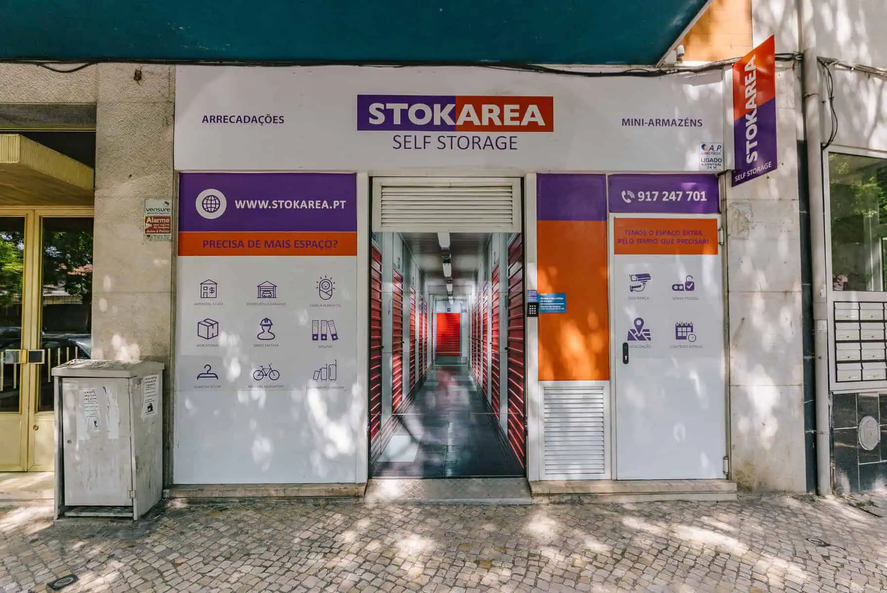 Stokarea - Mini Armazens Self storage Fachada de uma unidade de autoarmazenamento chamada Stokarea, com uma placa branca e roxa acima da entrada. As portas estão abertas, revelando um corredor alinhado com unidades de armazenamento vermelhas. As paredes externas exibem ícones e informações de contato. Lidar em Portugal