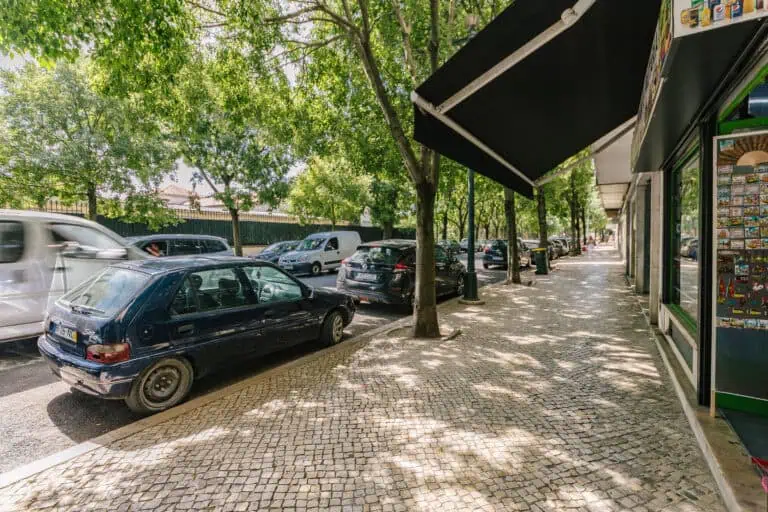 Stokarea - Mini Armazens Self storage Uma cena de rua com carros estacionados ao longo de uma calçada de paralelepípedos ladeada por árvores. Um carro preto está em primeiro plano, enquanto outros veículos e alguns pedestres são vistos mais abaixo na estrada. A cena é sombreada por um toldo à direita. Lidar em Portugal