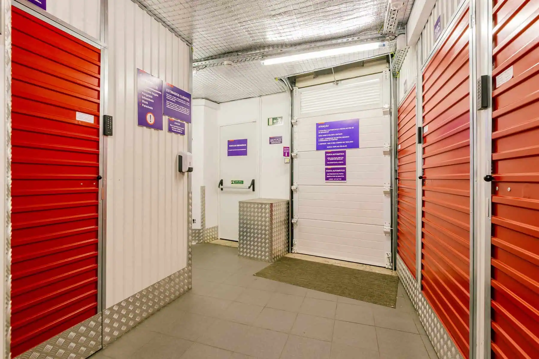 Stokarea - Mini Armazens Self storage Interior de um depósito com várias portas de enrolar laranja, teto branco e paredes com placas informativas. O piso é de ladrilho e há detalhes prateados nos rodapés. Lidar em Portugal
