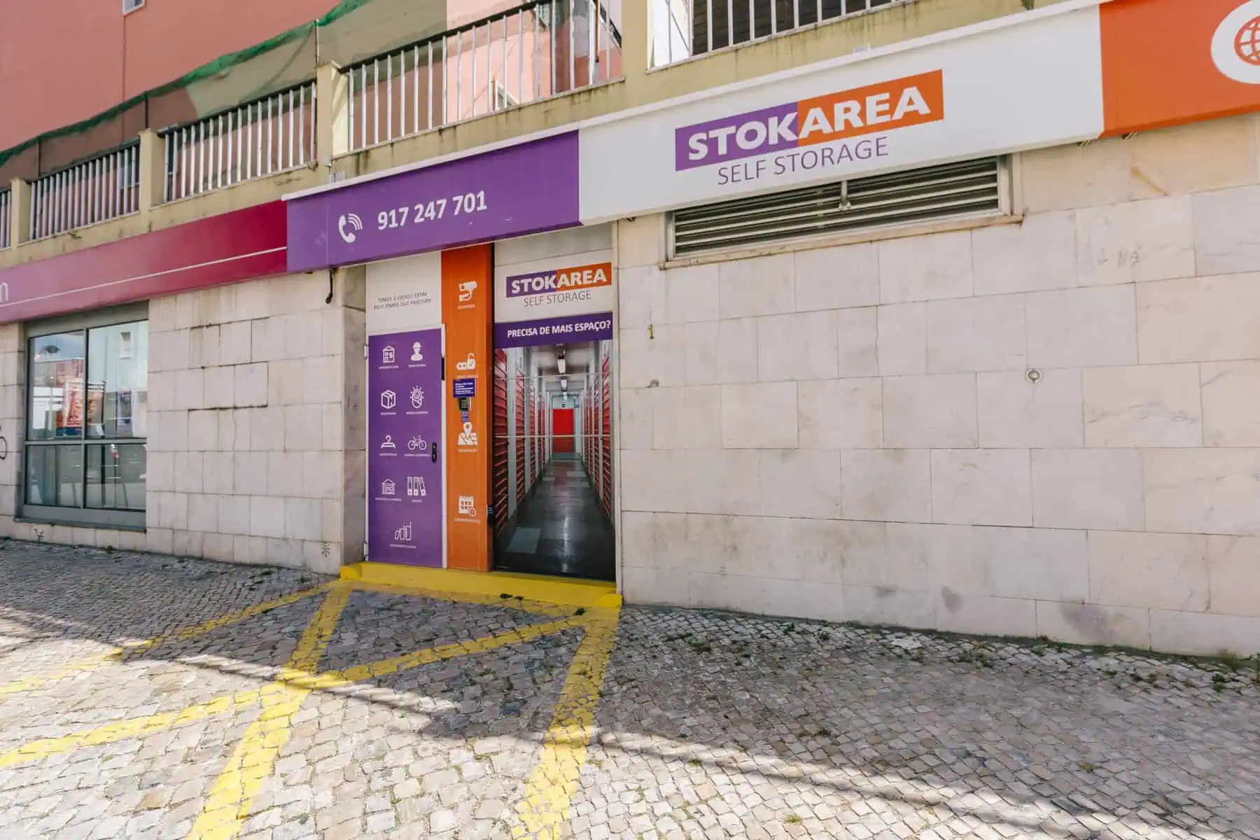 Stokarea - Mini Armazens Self storage Vista externa da entrada de uma unidade de autoarmazenamento STOKAREA, com uma fachada roxa e laranja com detalhes de contato e ícones de serviço na porta, em frente a uma passarela de paralelepípedos. Lidar em Portugal