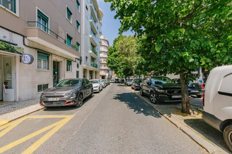 Stokarea - Mini Armazens Self storage Uma rua da cidade alinhada com carros estacionados em ambos os lados. Prédios e árvores alinham a rua, que se estende em direção à distância sob um céu azul claro. Uma marcação diagonal amarela é visível na estrada em primeiro plano. Lidar em Portugal