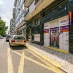 Stokarea - Mini Armazens Self storage Vista da rua com um carro bege estacionado em frente a um prédio rotulado Store Area. O prédio tem uma veneziana de metal e uma placa de fachada. Árvores se alinham no lado oposto da rua sob um céu limpo. Lidar em Portugal