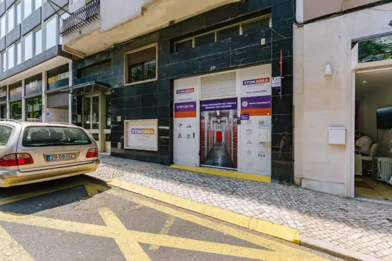 Stokarea - Mini Armazens Self storage Uma vista da rua de um edifício moderno com uma fachada de loja exibindo o nome STOKAREA. A entrada tem portas de vidro com sinalização, e um carro bege está estacionado próximo em uma rua de paralelepípedos com uma marcação de trânsito amarela. Lidar em Portugal