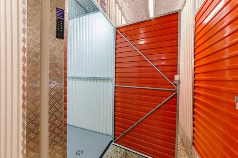 Stokarea - Mini Armazens Self storage Um corredor em um depósito com uma unidade aberta. As portas e paredes internas são laranja e branco brilhantes, e o piso é cinza salpicado. O teto tem uma malha de metal, e há iluminação visível de cima. Lidar em Portugal