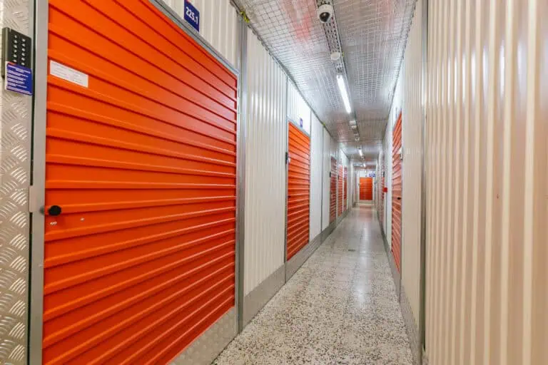 Stokarea - Mini Armazens Self storage Um corredor de depósito bem iluminado com portas de metal laranja em ambos os lados, levando a unidades de armazenamento individuais. O piso é salpicado, e as paredes e o teto são de cor clara, dando uma aparência limpa e organizada. Lidar em Portugal