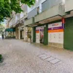 Stokarea - Mini Armazens Self storage Uma calçada ladeada por árvores e plantas corre ao longo de um prédio com paredes verdes. Há um escritório imobiliário com placas e uma caixa de correio à direita. Uma pessoa caminha ao longe perto de uma rua coberta por uma copa de árvores. Lidar em Portugal