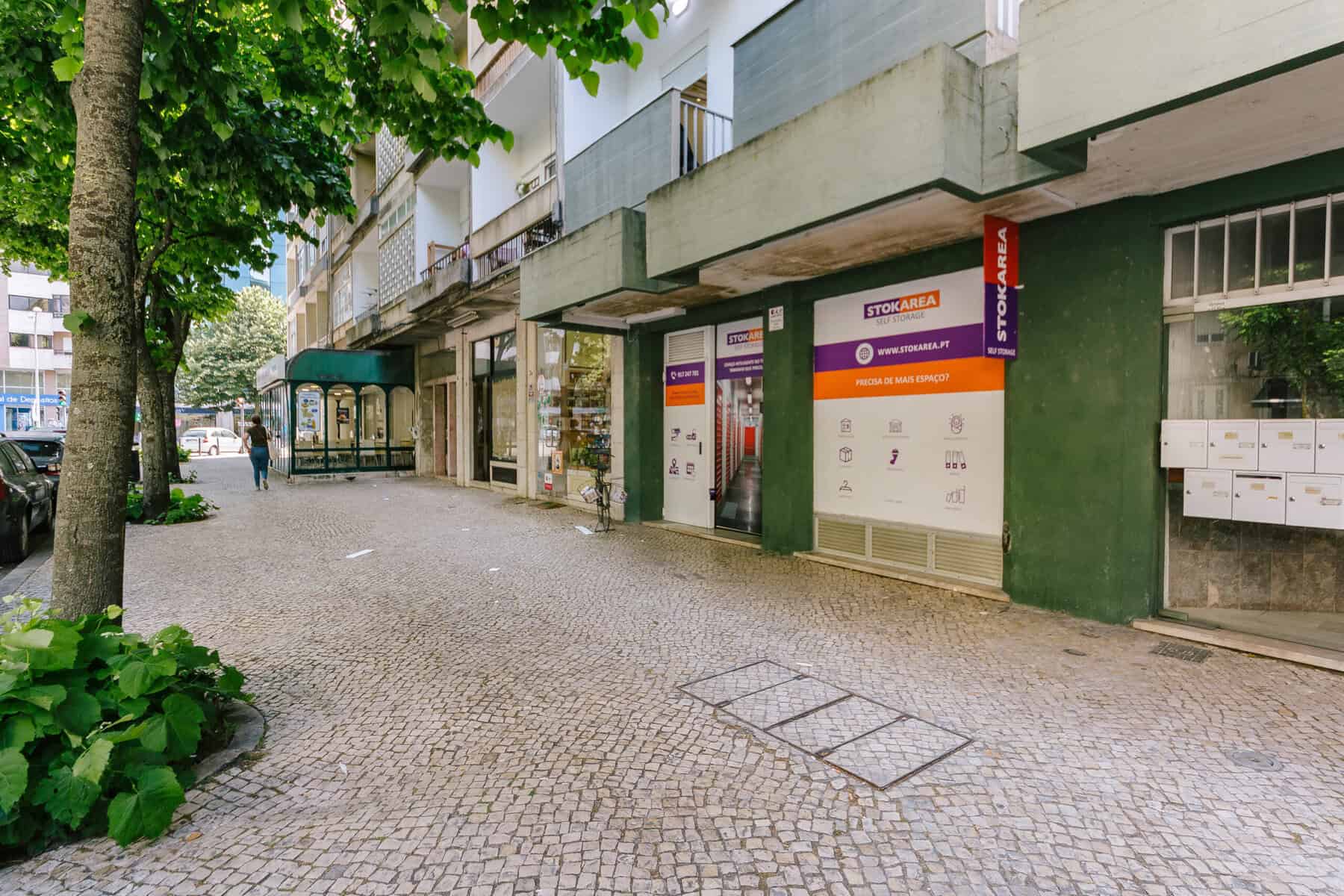 Stokarea - Mini Armazens Self storage Uma calçada ladeada por árvores e plantas corre ao longo de um prédio com paredes verdes. Há um escritório imobiliário com placas e uma caixa de correio à direita. Uma pessoa caminha ao longe perto de uma rua coberta por uma copa de árvores. Lidar em Portugal