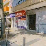 Stokarea - Mini Armazens Self storage Uma vitrine com um dossel laranja e vermelho exibindo STOKAREA em uma rua ensolarada. Fica ao lado de um prédio cinza com uma árvore e veículos estacionados nas proximidades. A calçada é pavimentada com paralelepípedos. Lidar em Portugal