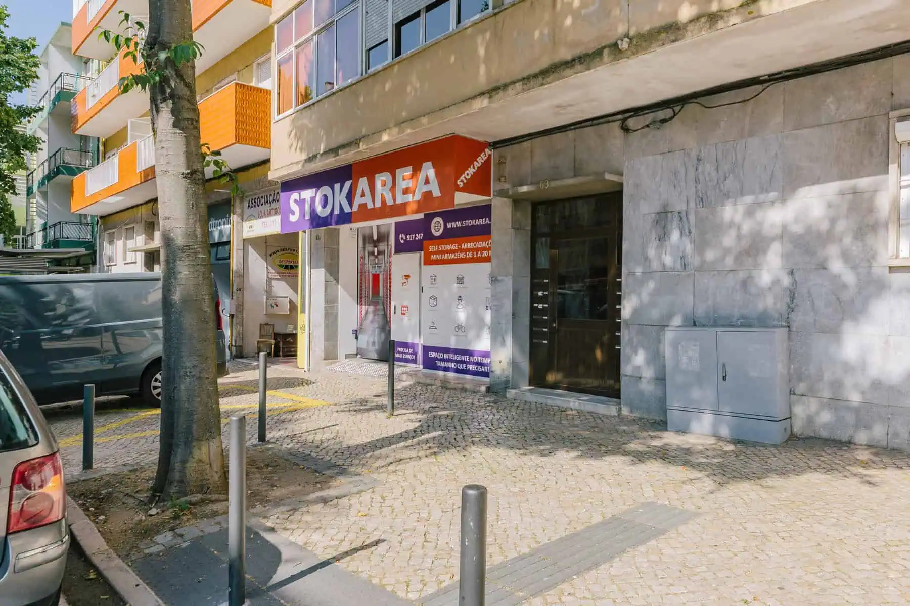 Stokarea - Mini Armazens Self storage Uma vitrine com um dossel laranja e vermelho exibindo STOKAREA em uma rua ensolarada. Fica ao lado de um prédio cinza com uma árvore e veículos estacionados nas proximidades. A calçada é pavimentada com paralelepípedos. Lidar em Portugal