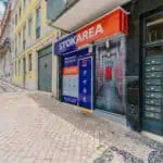 Stokarea - Mini Armazens Self storage Vista da rua de um self-storage com uma placa azul e laranja dizendo STOKAREA. A entrada fica em uma calçada de paralelepípedos ao lado de um prédio com paredes amarelas. Várias caixas de correio são visíveis à direita da entrada. Lidar em Portugal