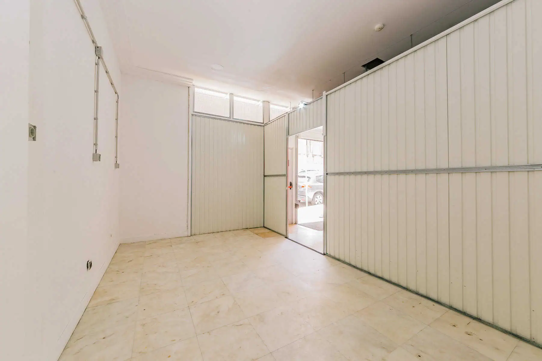 Stokarea - Mini Armazens Self storage Uma sala vazia e iluminada com piso de ladrilho e paredes de painéis brancos. Há uma porta de correr parcialmente aberta que leva para fora, permitindo a entrada de luz natural. O espaço parece ser uma garagem ou área de armazenamento. Lidar em Portugal