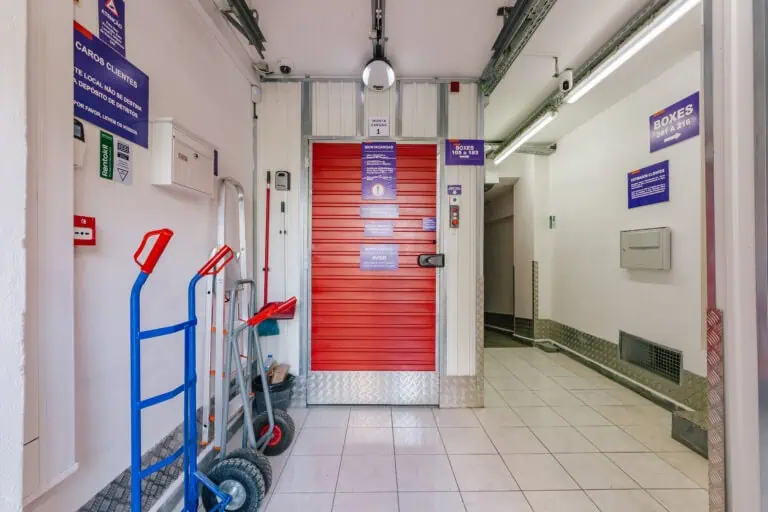 Stokarea - Mini Armazens Self storage O Stokarea Self Storage oferece mini armazéns para alugar, com uma porta de veneziana vermelha fechada e cercado por equipamentos como carrinhos de mão e um carrinho. Instruções claras de segurança são postadas nas paredes de azulejos bem iluminadas, garantindo uma experiência de armazenamento eficiente e segura. Lidar em Portugal