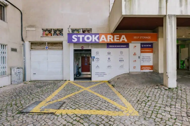 Stokarea - Mini Armazens Self storage A imagem captura a entrada do Stokarea, um self-storage com mini armazéns para alugar. Uma vibrante placa roxa e laranja exibe ícones de armazenamento acima. O primeiro plano de paralelepípedos apresenta um triângulo amarelo marcado, dando boas-vindas aos clientes ao espaço organizado dentro. Lidar em Portugal
