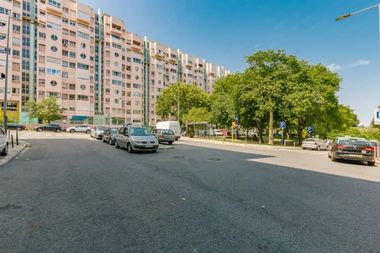 Stokarea - Mini Armazens Self storage Uma cena de rua com carros estacionados ao longo da calçada, uma van branca e um aglomerado de árvores. Ao fundo, a unidade de armazenamento Stokarea Self Storage se destaca ao lado de um grande edifício residencial rosa sob um céu azul claro, oferecendo mini armazéns para alugar. Lidar em Portugal