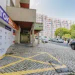 Stokarea - Mini Armazens Self storage Cena de rua urbana com fachadas de lojas, com um edifício exibindo a placa "Stokarea Self Storage - mini armazéns para alugar". Um carro está estacionado na estrada de paralelepípedos, enquanto apartamentos altos e árvores estão sob um céu azul. Lidar em Portugal