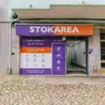 Stokarea - Mini Armazens Self storage A fachada da Stokarea Self Storage apresenta uma placa roxa e laranja. A entrada fechada é emoldurada por texto informativo e ícones, convidando você a explorar seus mini armazéns para alugar. Abaixo, a calçada ostenta azulejos com padrões intrincados. Lidar em Portugal