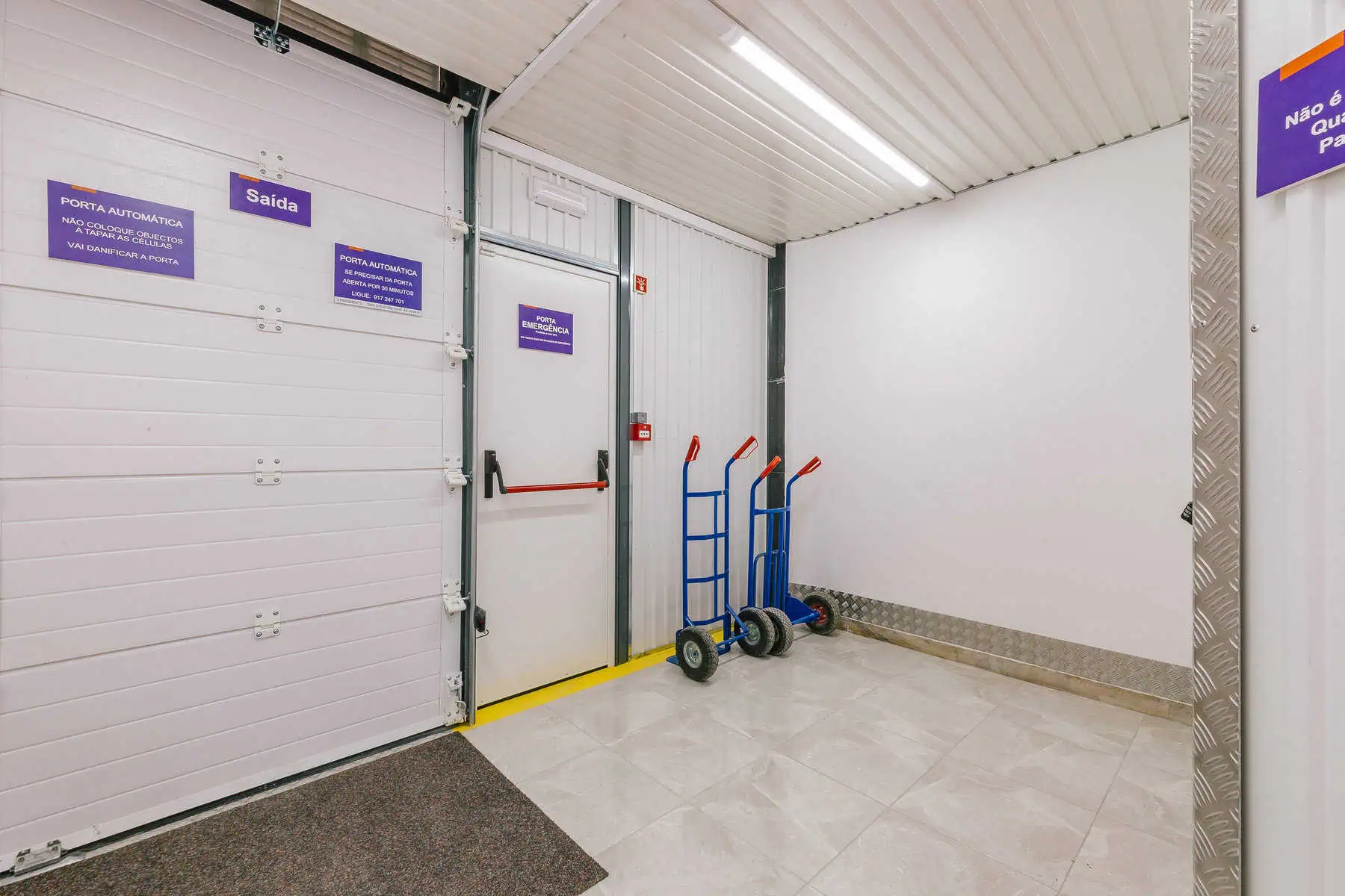 Stokarea - Mini Armazens Self storage Um corredor com uma porta branca rotulada Saída à esquerda, cercada por placas de segurança, indica o comprometimento da Stokarea Self Storage com a segurança. Perto dali, dois carrinhos de mão estão prontos. O corredor apresenta piso e paredes de ladrilhos de cor clara, criando um espaço iluminado e eficiente, ideal para seus mini armazéns para alugar. Lidar em Portugal