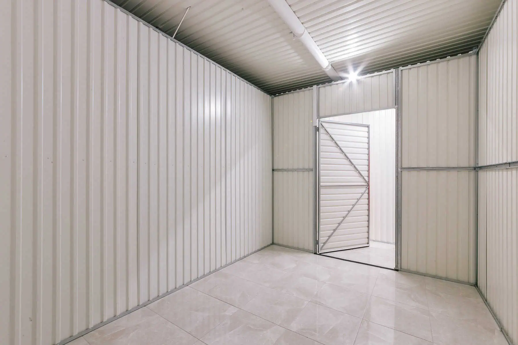 Stokarea - Mini Armazens Self storage Uma sala vazia com paredes de metal corrugado branco e um piso de ladrilho de cor clara. A porta, feita de metal corrugado semelhante, está ligeiramente aberta, e uma iluminação brilhante ilumina o espaço de cima. Perfeito para as opções de mini armazéns para alugar da Stokarea Self Storage. Lidar em Portugal