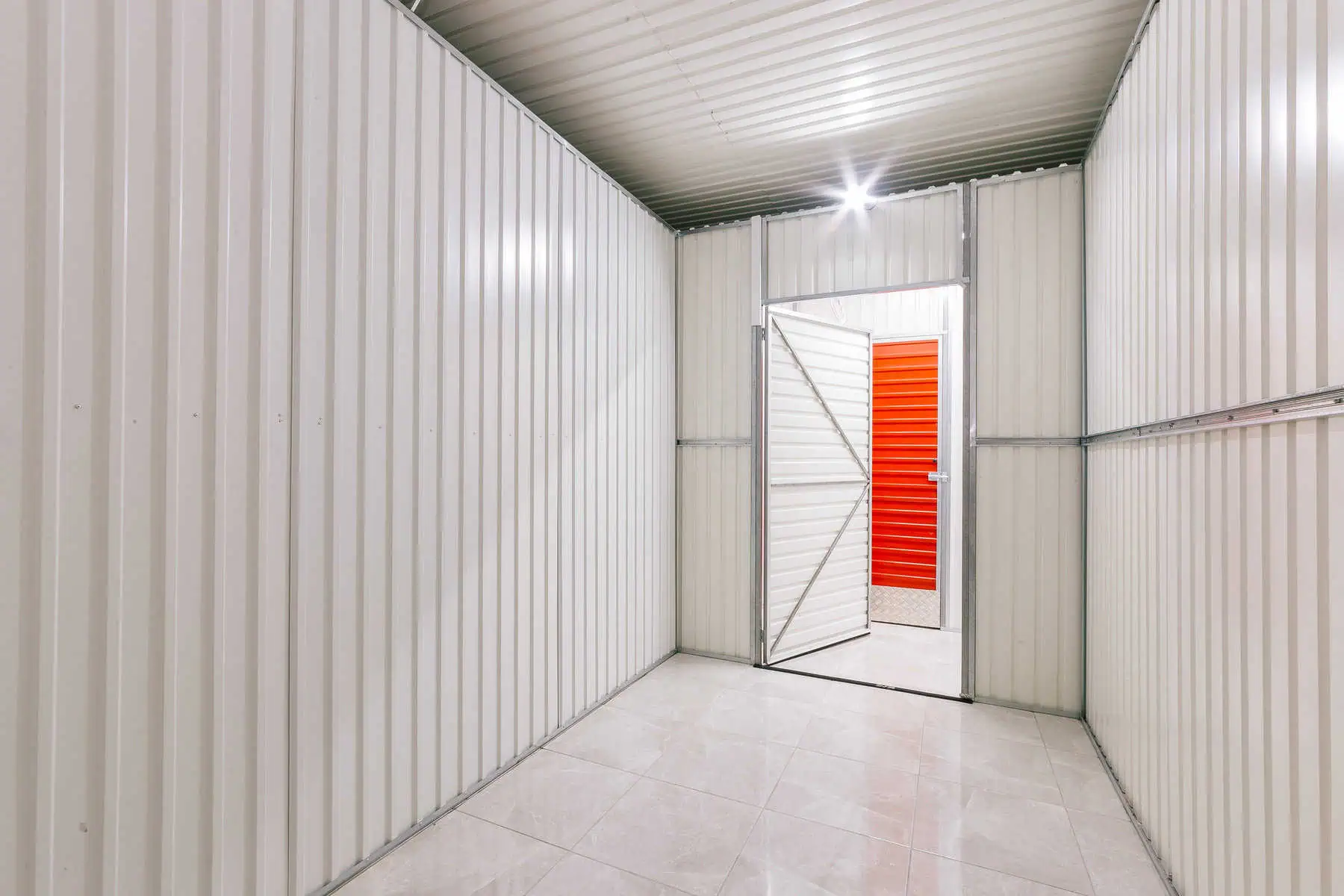 Stokarea - Mini Armazens Self storage A unidade de Self Storage Stokarea brilhante apresenta paredes e teto de metal corrugado branco, piso de cerâmica e uma porta aberta que leva a um corredor com uma impressionante porta vermelha à vista. Perfeito para quem busca mini armazéns para alugar. Lidar em Portugal