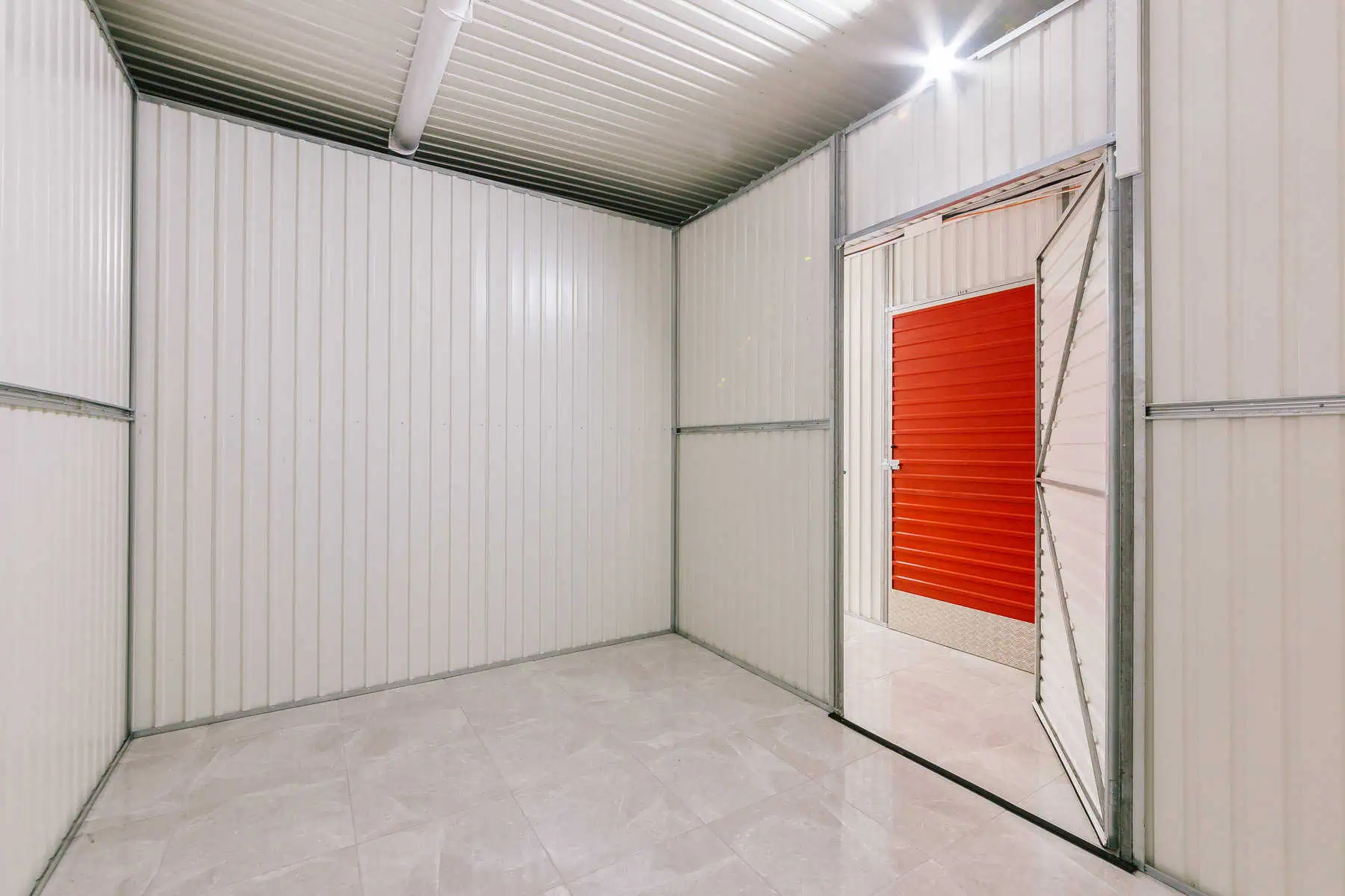 Stokarea - Mini Armazens Self storage A unidade de armazenamento vazia, parte do Stokarea Self Storage, apresenta paredes de metal branco e piso de cerâmica. Sua porta aberta revela uma parede externa vermelha. Luzes de teto fornecem iluminação brilhante, ideal para aqueles que buscam mini armazéns para alugar. Lidar em Portugal