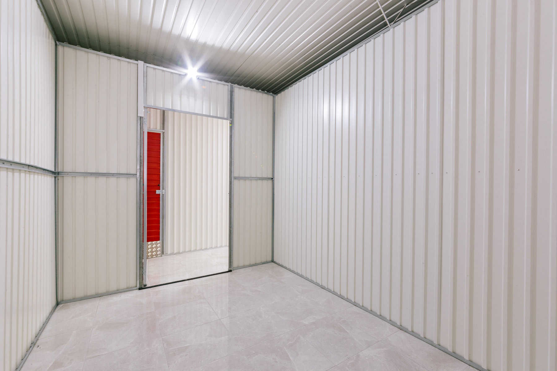 Stokarea - Mini Armazens Self storage O interior desta unidade de self-storage Stokarea apresenta paredes de metal corrugado de cor clara e piso de cerâmica. A porta aberta revela uma parede vermelha em uma unidade adjacente, enquanto a iluminação brilhante realça a aparência limpa e espaçosa, ideal para quem busca mini armazéns para alugar. Lidar em Portugal