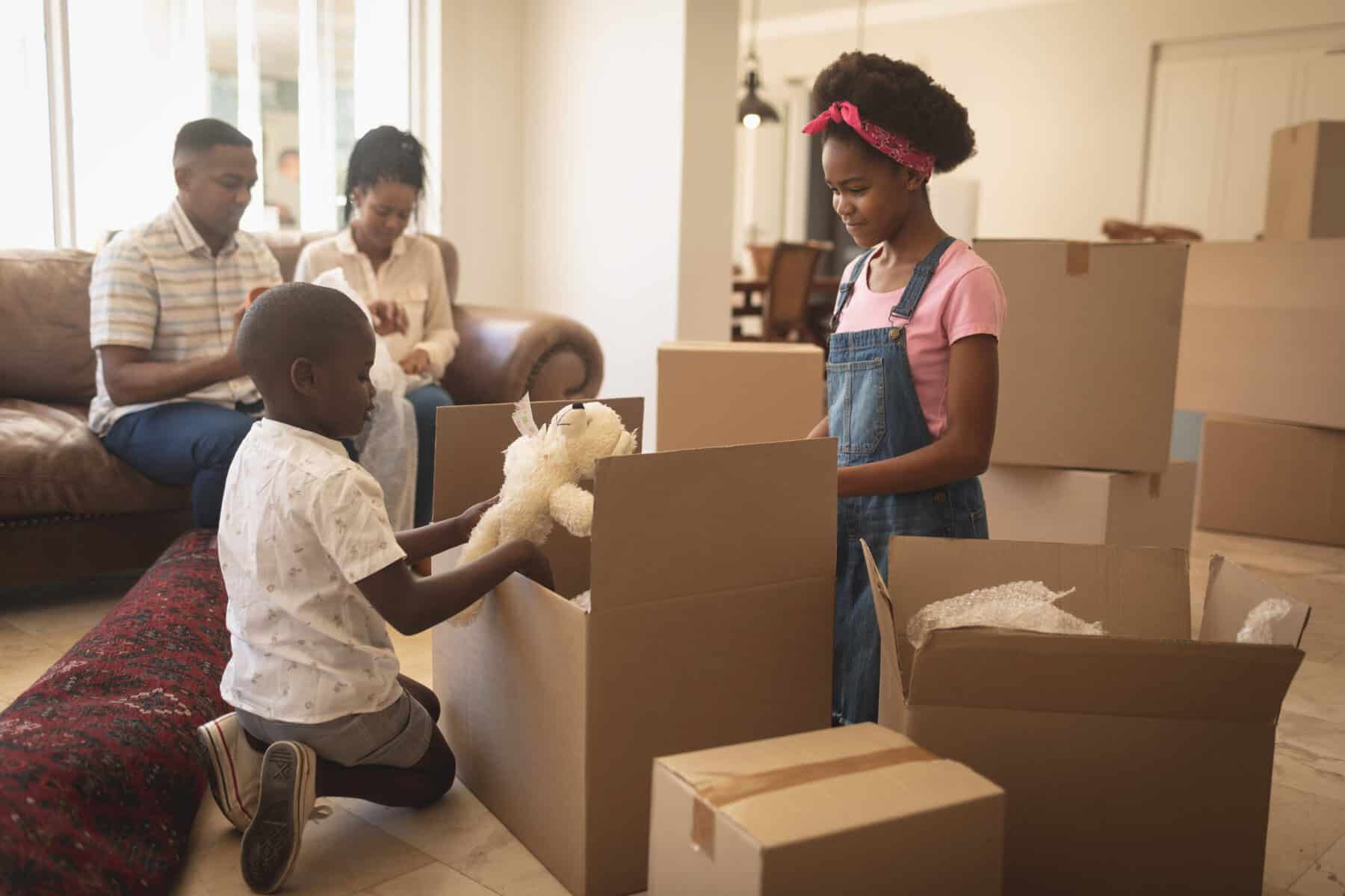 Stokarea - Mini Armazens Self storage Uma família está ocupada desempacotando caixas de mudança na sala de estar. Uma jovem garota de macacão e faixa na cabeça abre animadamente uma caixa, enquanto seu irmão admira um ursinho de pelúcia. Dois adultos sentam-se em um sofá próximo, cercados por caixas de sua aventura de self storage - mini armazéns para alugar. Lidar em Portugal