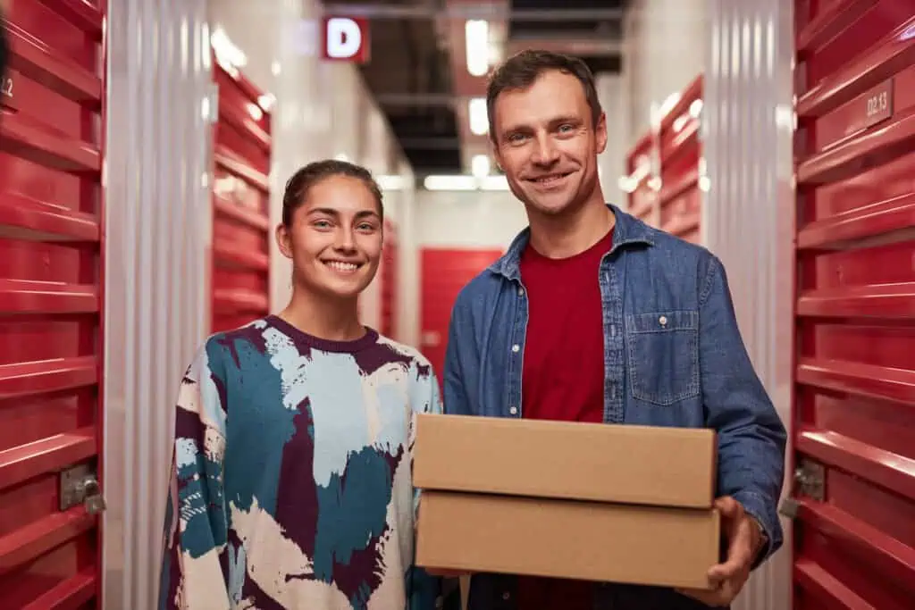 Stokarea - Mini Armazens Self storage Um homem e uma mulher estão em um corredor de Self Storage da Stokarea, sorrindo. O homem segura duas caixas de papelão. As unidades de armazenamento ao redor deles têm portas vermelhas, exibindo a opção perfeita de mini armazéns para alugar, com uma luz de teto iluminando o fundo. Lidar em Portugal
