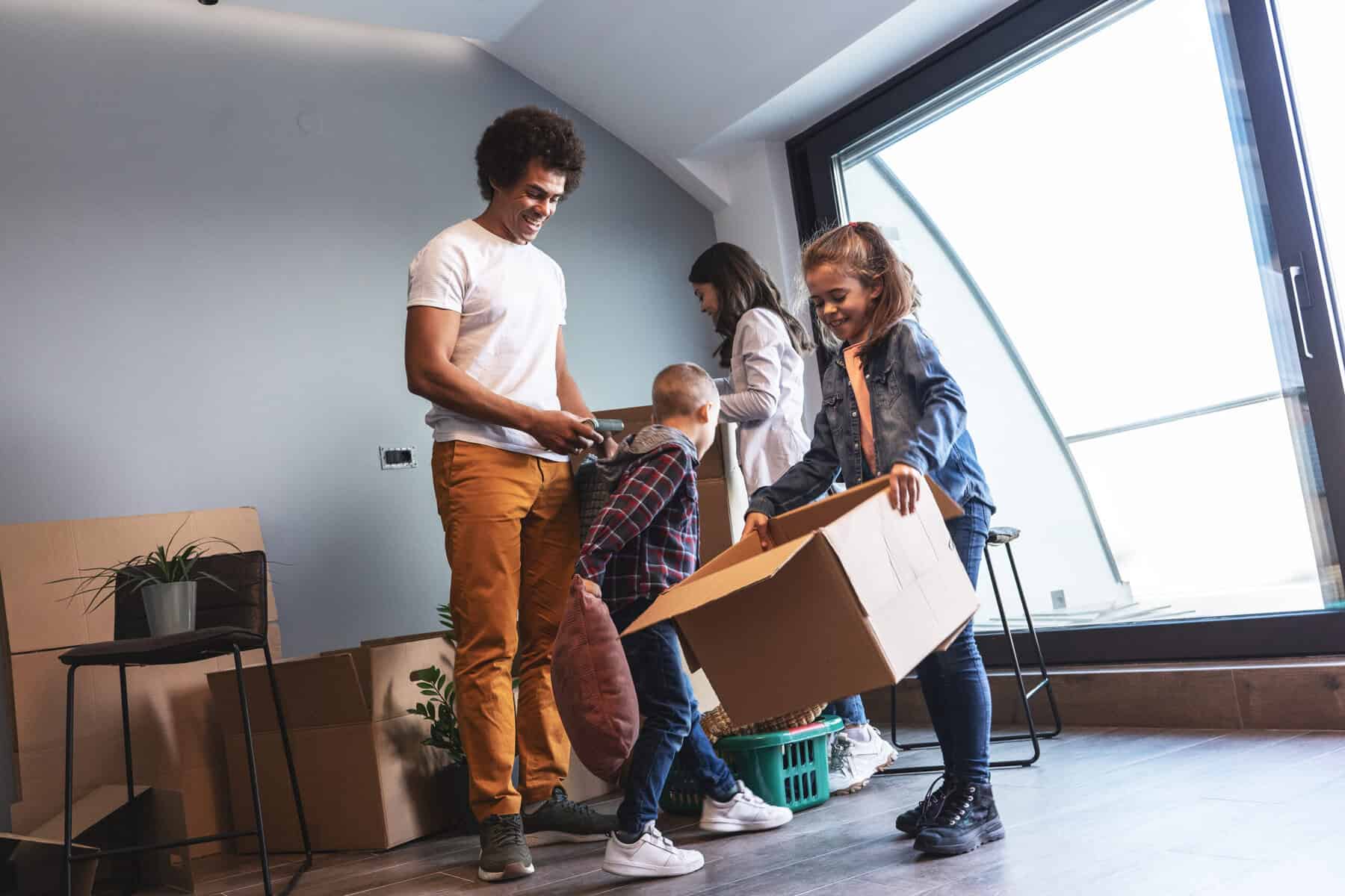 Stokarea - Mini Armazens Self storage Uma família está dentro de uma sala com janelas grandes, desempacotando caixas do Stokarea Self Storage. O pai e a mãe estão sorrindo, enquanto seus dois filhos ajudam carregando uma caixa de papelão. Várias caixas e um vaso de plantas os cercam em seus mini armazéns para alugar. Lidar em Portugal