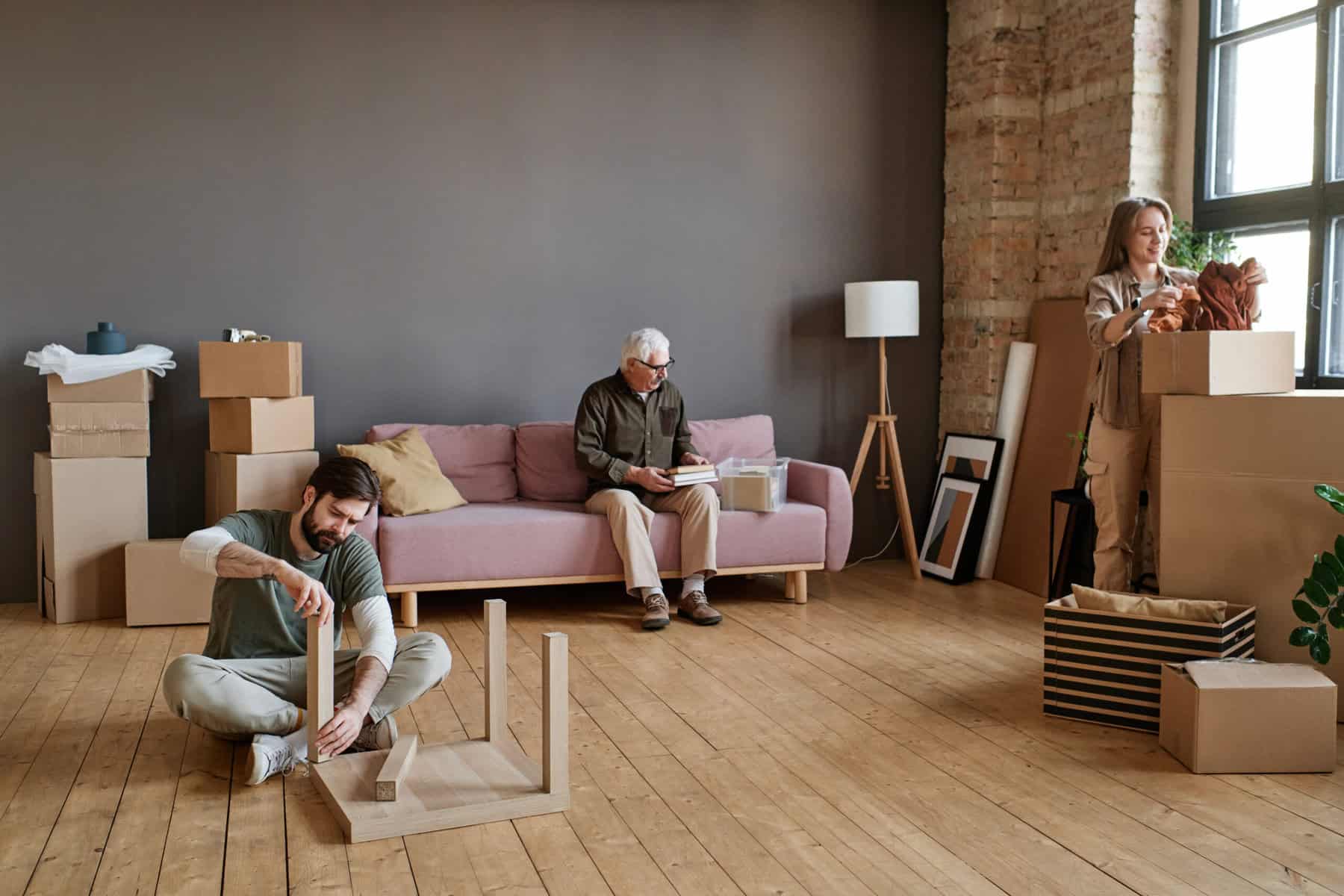 Stokarea - Mini Armazens Self storage Em uma sala espaçosa com piso de madeira, um homem monta móveis no chão, enquanto um homem mais velho lê uma revista em um sofá rosa. Perto dali, uma mulher desempacota uma caixa cercada por caixas de mudança e itens espalhados. Eles são gratos pelo espaço extra fornecido pela Stokarea Self Storage - mini armazéns para alugar. Lidar em Portugal