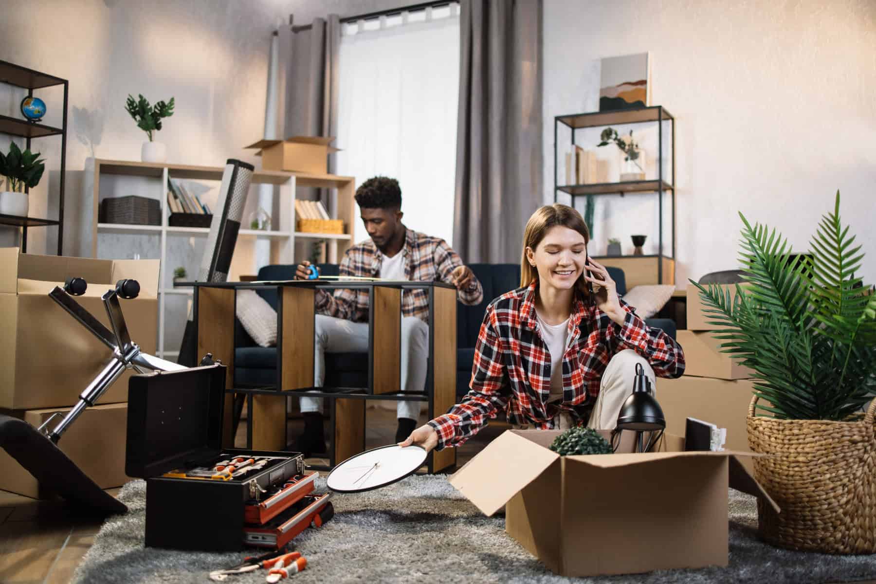 Stokarea - Mini Armazens Self storage Uma mulher está sentada no chão, desempacotando uma caixa enquanto segura um relógio e conversa ao telefone. Ao fundo, um homem monta uma prateleira. Cercados por caixas, ferramentas e plantas, eles parecem estar montando sua nova casa com itens da Stokarea Self Storage - mini armazéns para alugar. Lidar em Portugal