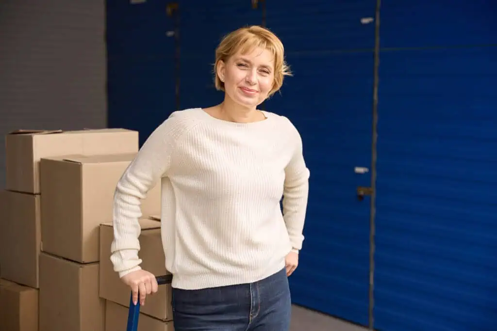 Stokarea - Mini Armazens Self storage Uma pessoa com cabelo curto está sorrindo, vestindo um suéter branco e jeans. Ela está em frente a caixas de papelão empilhadas e portas azuis de unidades de armazenamento no Stokarea Self Storage, segurando um carrinho para mover itens de seus mini armazéns para alugar. Lidar em Portugal
