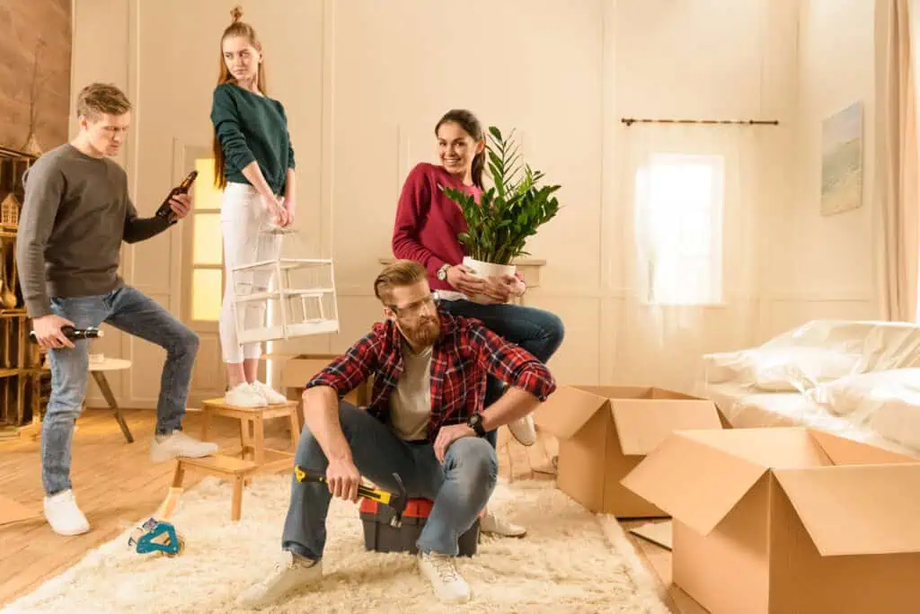 Stokarea - Mini Armazens Self storage Em uma sala aconchegante e movimentada, quatro pessoas navegam por caixas de mudança. Uma se ajoelha com uma caixa de ferramentas, outra embala cuidadosamente uma planta, enquanto alguém fica em pé em um banquinho ajustando uma pequena prateleira, e a última pessoa segura uma garrafa em meio à transformação — ideal para quem está considerando o Stokarea Self Storage. Lidar em Portugal