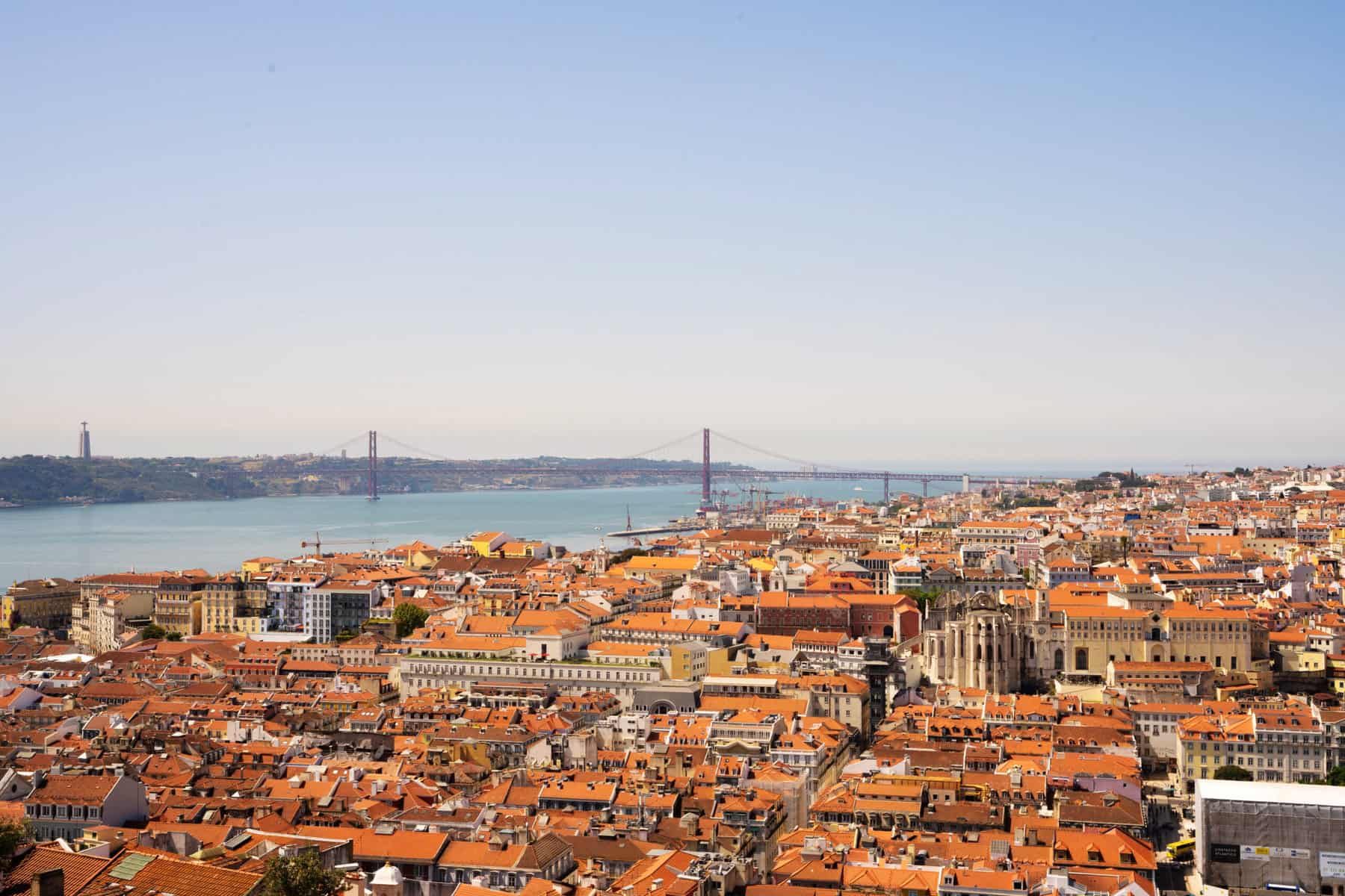 Stokarea - Mini Armazens Self storage Uma vista panorâmica de Lisboa, Portugal, apresentando uma paisagem pontilhada com prédios de telhado vermelho. O Rio Tejo e a proeminente Ponte 25 de Abril são visíveis ao fundo sob um céu azul claro. Lidar em Portugal