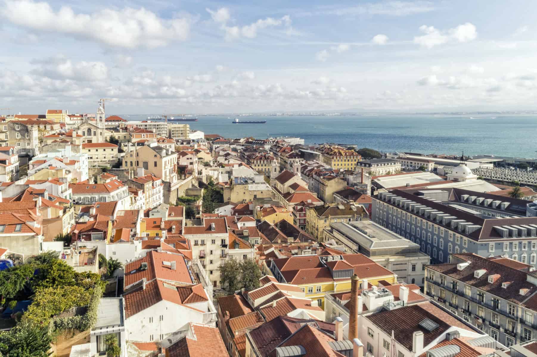 Stokarea - Mini Armazens Self storage Vista aérea de uma cidade costeira com uma mistura de prédios tradicionais de telhado vermelho e estruturas modernas. A cidade é cercada por um corpo dágua, com um navio visível à distância e céu parcialmente nublado acima. Lidar em Portugal