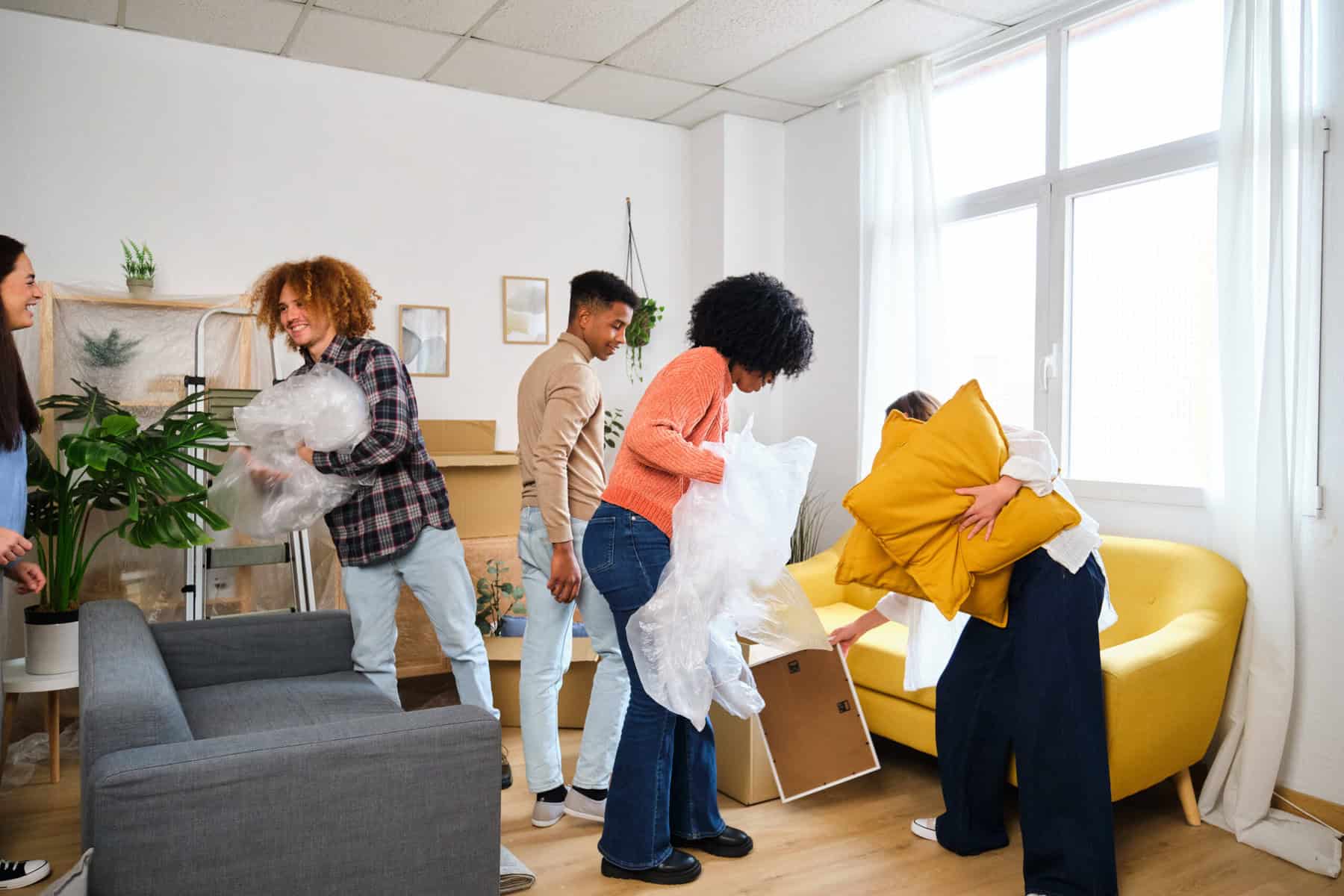 Stokarea - Mini Armazens Self storage Quatro pessoas estão desempacotando coisas em uma sala de estar iluminada. Uma pessoa está segurando plástico-bolha, outra está carregando almofadas amarelas e uma terceira está manuseando um porta-retratos. Um sofá cinza e uma cadeira amarela estão na sala, com plantas decorando o espaço. Lidar em Portugal