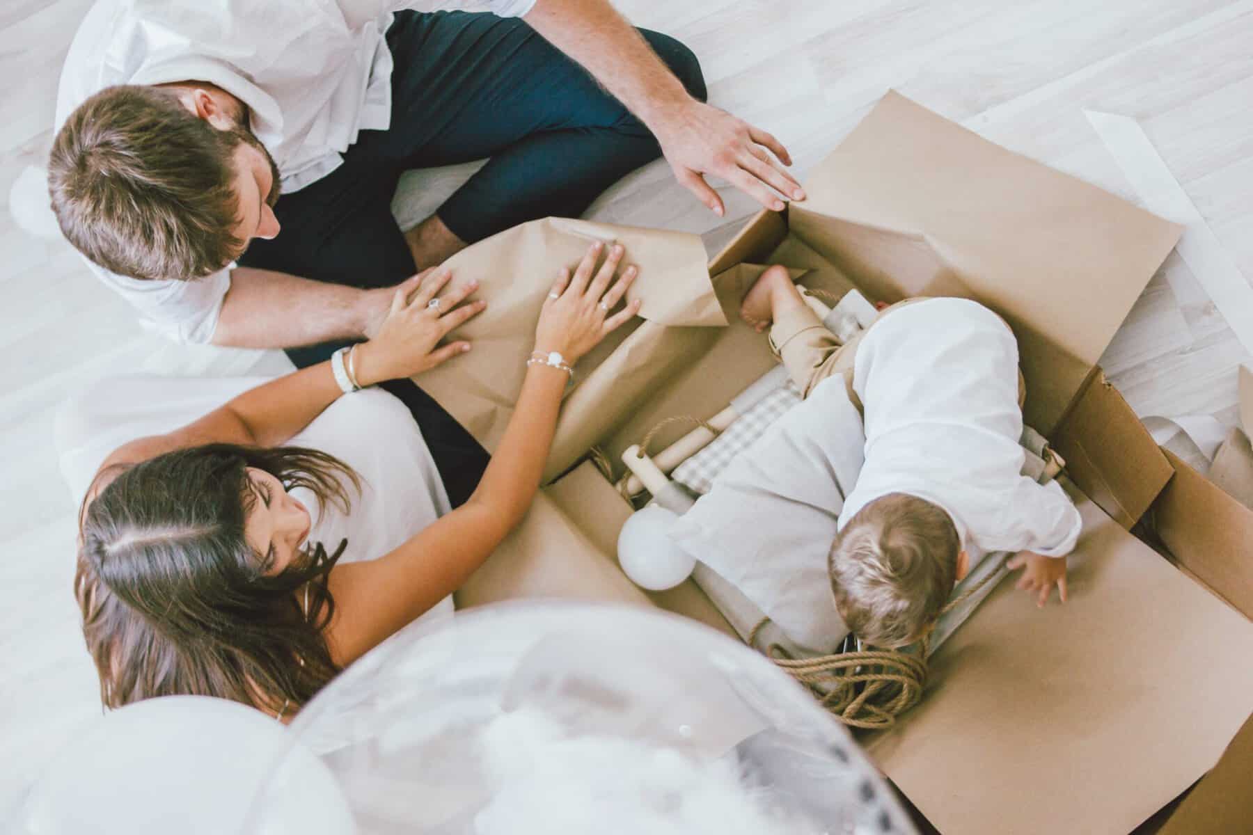 Stokarea - Mini Armazens Self storage Uma família está sentada no chão abrindo uma caixa de papelão. Um homem e uma mulher adultos ajudam uma criança pequena que está engatinhando em cima da caixa. A cena sugere um momento de exploração ou desempacotamento juntos. Lidar em Portugal