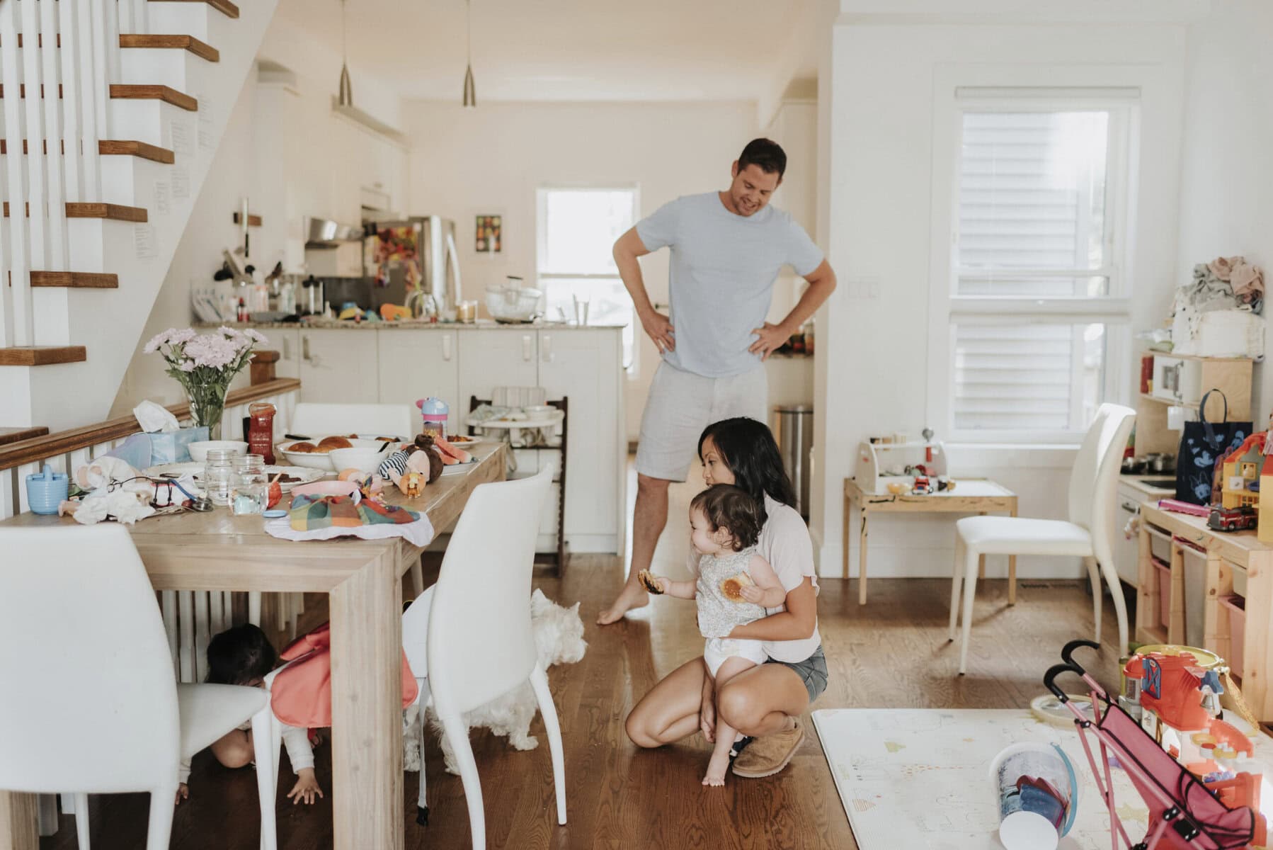 Stokarea - Mini Armazens Self storage Uma mãe ajoelha-se no chão, segurando um bebê, enquanto um homem está perto com as mãos nos quadris, sorrindo. Uma criança espia por baixo de uma mesa, e um cachorro está por perto. O quarto está abarrotado de brinquedos e itens domésticos, criando uma cena familiar animada. Lidar em Portugal