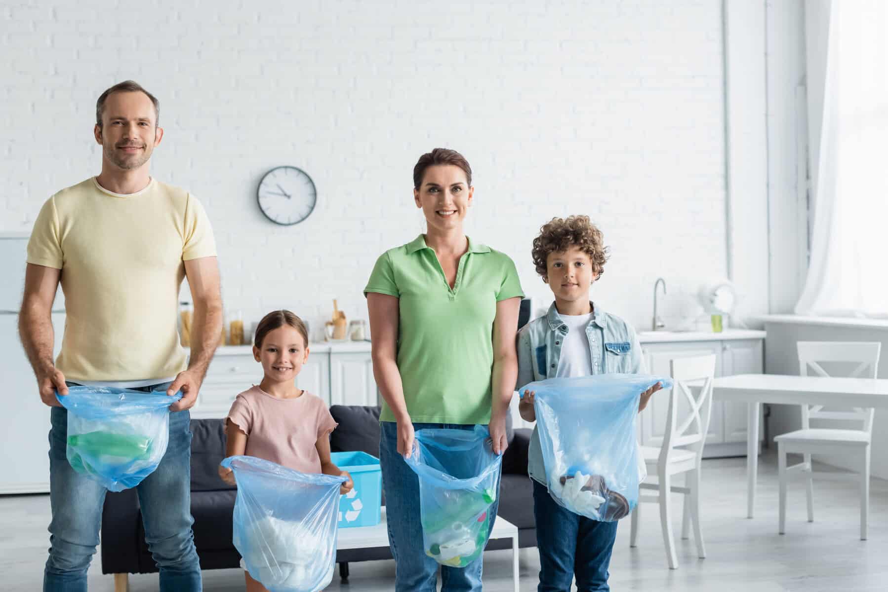 Stokarea - Mini Armazens Self storage Uma família de quatro pessoas, incluindo um homem adulto, uma mulher, uma menina e um menino, está em uma cozinha iluminada segurando sacolas azuis de reciclagem cheias de recicláveis variados, sorrindo para a câmera. Um relógio é visível na parede de tijolos brancos atrás deles. Lidar em Portugal