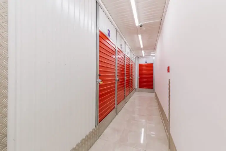 Stokarea - Mini Armazens Self storage Um longo corredor com paredes e teto brancos, com portas vermelhas de unidade de armazenamento à esquerda. O piso é de ladrilhos de cor clara, e luzes fluorescentes iluminam o corredor. Há uma pequena caixa vermelha de alarme de incêndio na parede direita. Lidar em Portugal