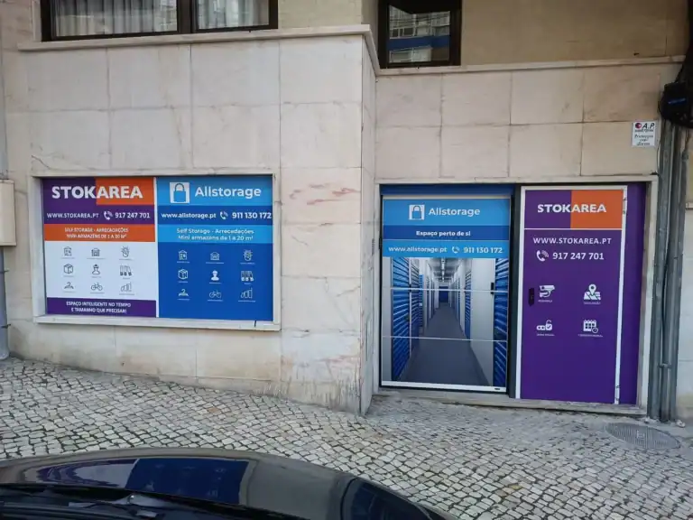 Stokarea - Mini Armazens Self storage Exterior de um edifício com sinalização para Stokarea e Allstorage. A placa da esquerda exibe informações de contato e serviços. A entrada da direita mostra um corredor que leva a unidades de armazenamento. O chão é pavimentado com paralelepípedos. Lidar em Portugal