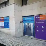 Stokarea - Mini Armazens Self storage Vitrine com dois negócios adjacentes. A esquerda apresenta uma placa azul Allstorage com texto e ícones brancos. A direita exibe uma placa roxa Stokarea com informações de contato. Um caminho de paralelepípedos e parte de um edifício são visíveis. Lidar em Portugal