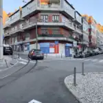 Stokarea - Mini Armazens Self storage Um prédio de esquina com tijolos vermelhos e varandas fica em um cruzamento com trilhos de bonde, carros estacionados, scooters e calçadas de paralelepípedos em um dia claro. As placas e as vitrines das lojas são visíveis no nível da rua. Lidar em Portugal